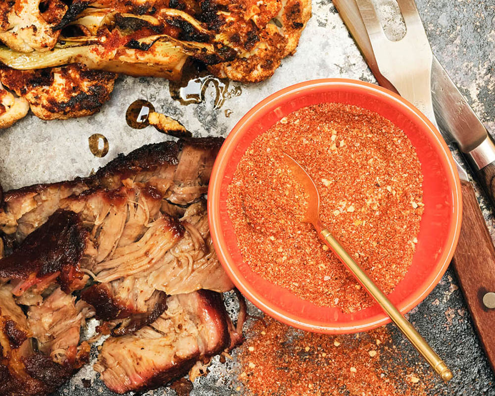 Red dry spice rub in a bowl on a tray with cooked meat and vegetables.