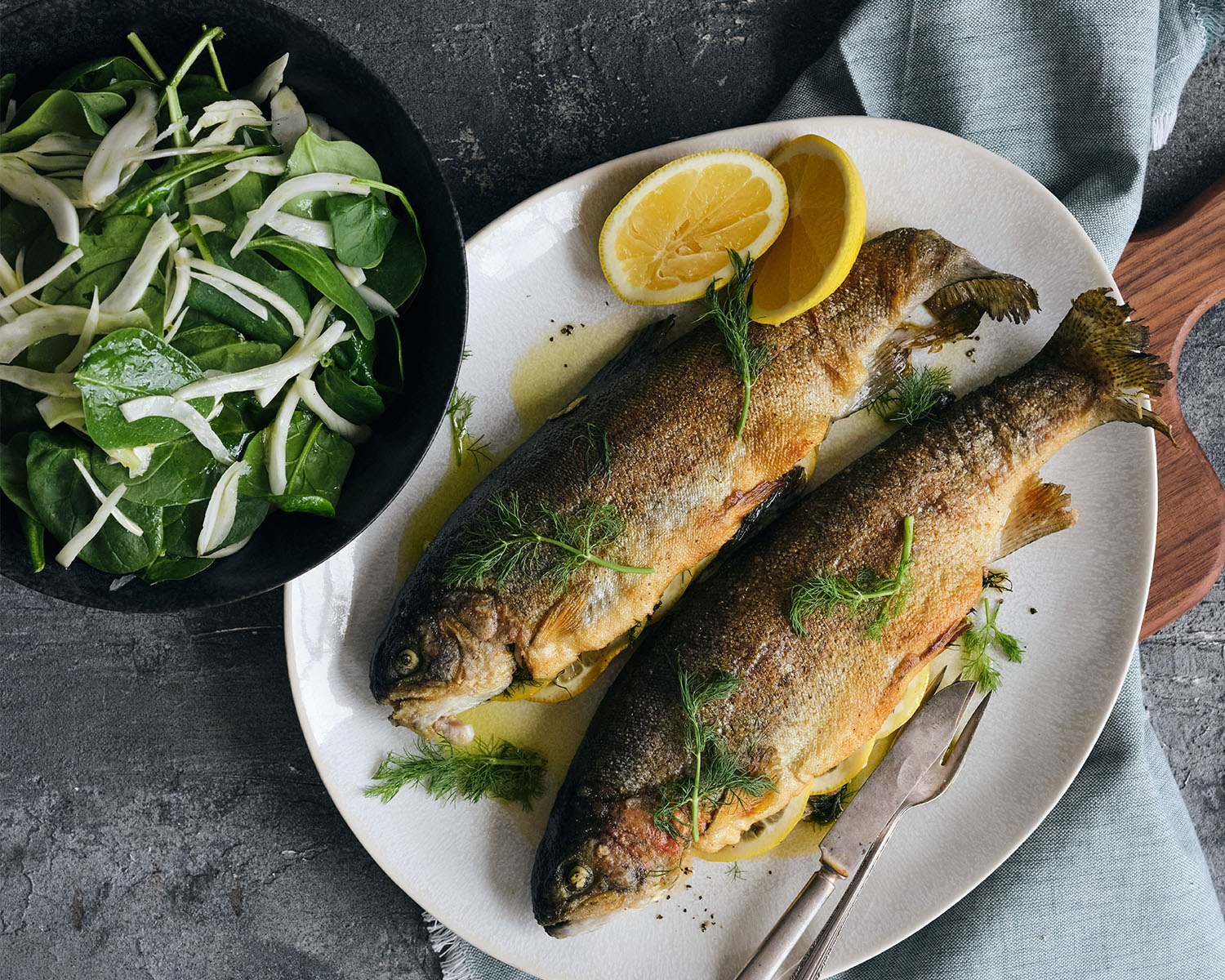 two cooked whole trout on a plate with lemon slices and a green salad