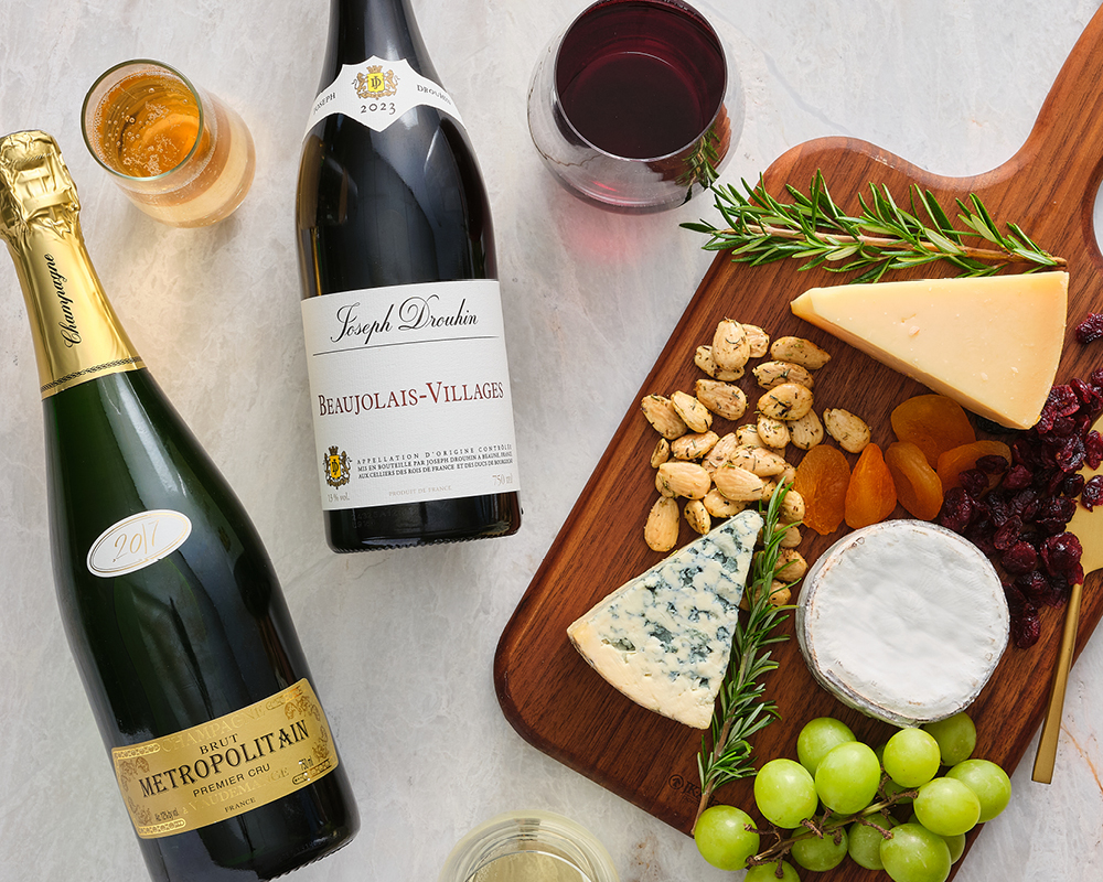 Metropolitain Champagne and Joseph Broughin Beaujolais-Villages Wine next to assorted cheese and dried fruit and nuts charcuterie board