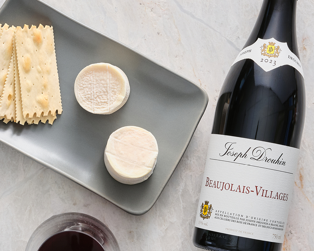 Bijou Chevre Cheese next to crackers on a gray stone setting plate with a bottle of Joseph Drouhin Beaujolais-Villages red wine on marble table