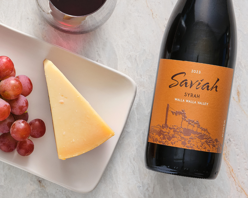 Ewephoria Gouda Cheese next to grapes on white plate next to a glass of wine and a Saviah Syrah Red Wine Bottle to the right all on top of marble table