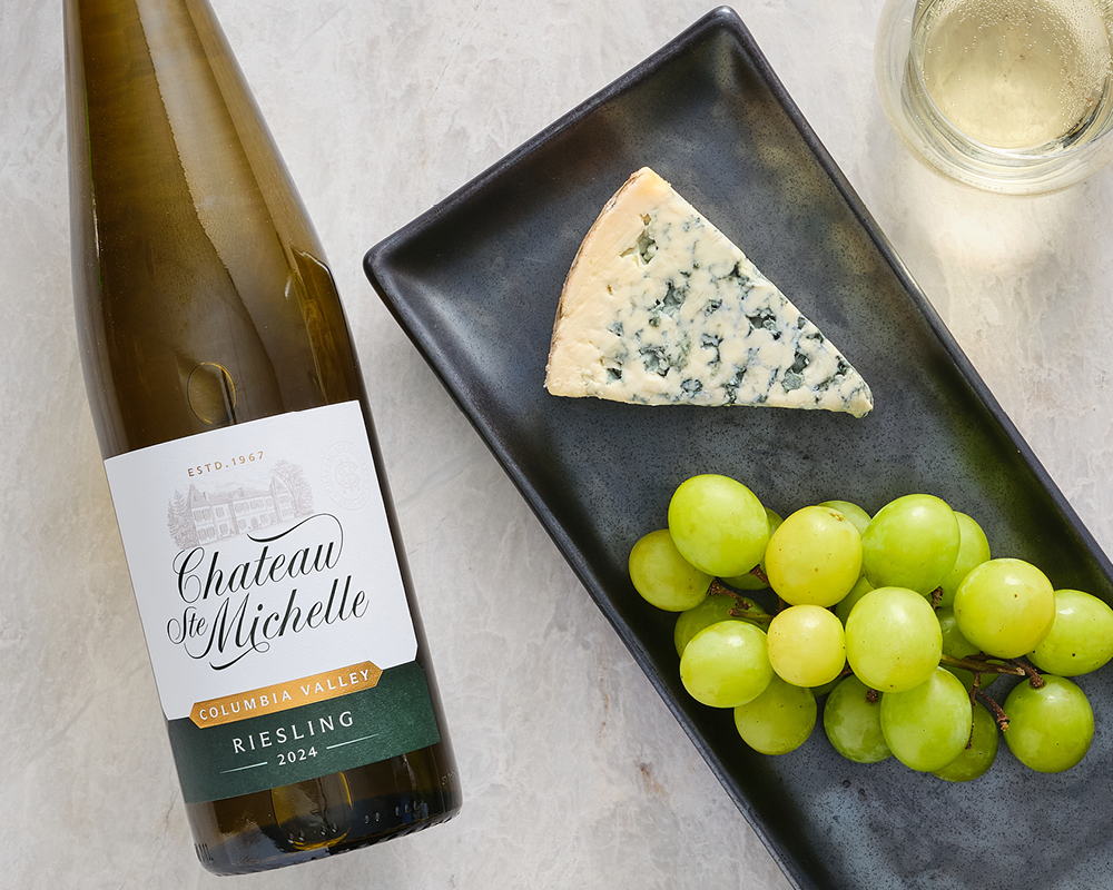 Blue d'Auvergne Cheese next to green grapes on black stone serving platter with glass or white wine to the right and a bottle of Chateau Ste. Michelle Harvest Select Riesling White Wine Bottle to the left on marble table