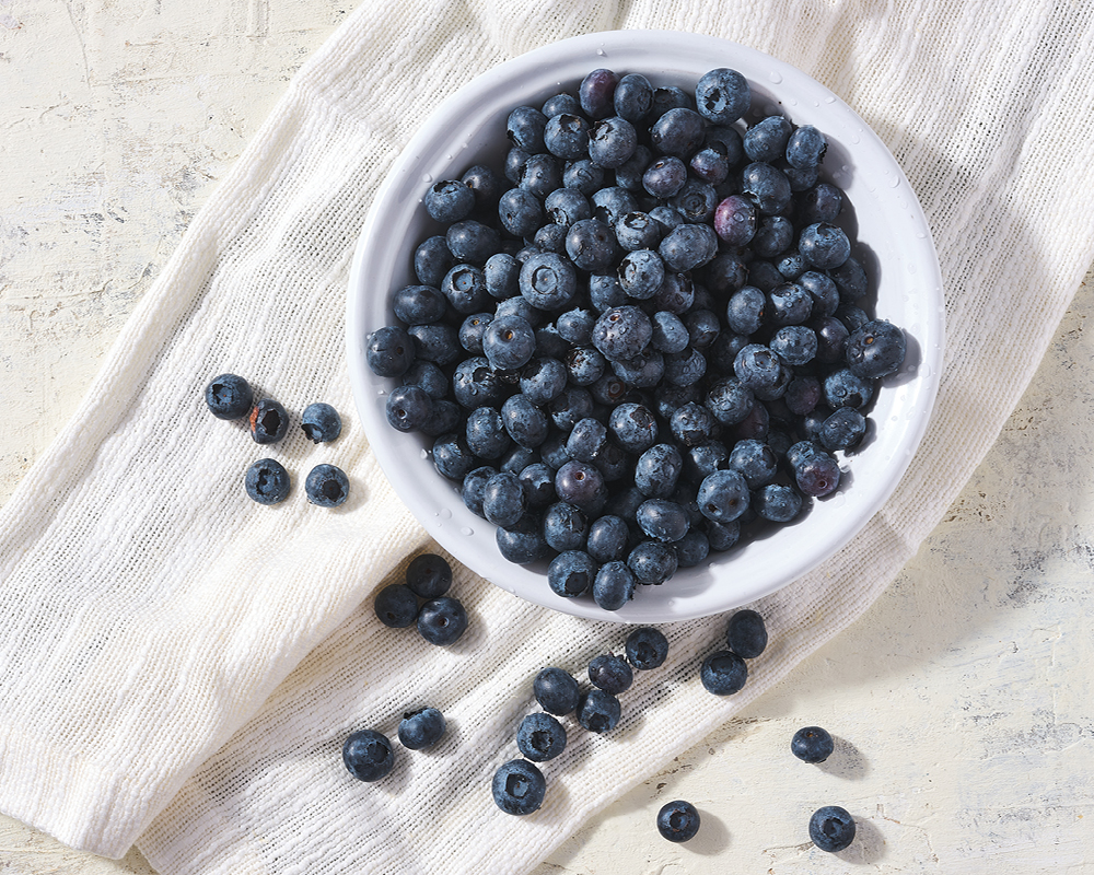 Blueberries in a white bowl with blueberries scattered on white linen