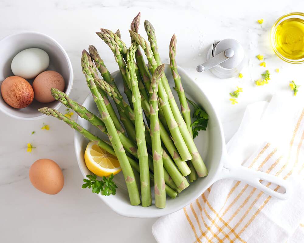 asparagus stalks in a white pan with a lemon half and stalks of parsley. a bowl of white and brown eggs sits to the left side. a peppermill, loose flowers, a dish of olive oil and a dish towel are on the right..