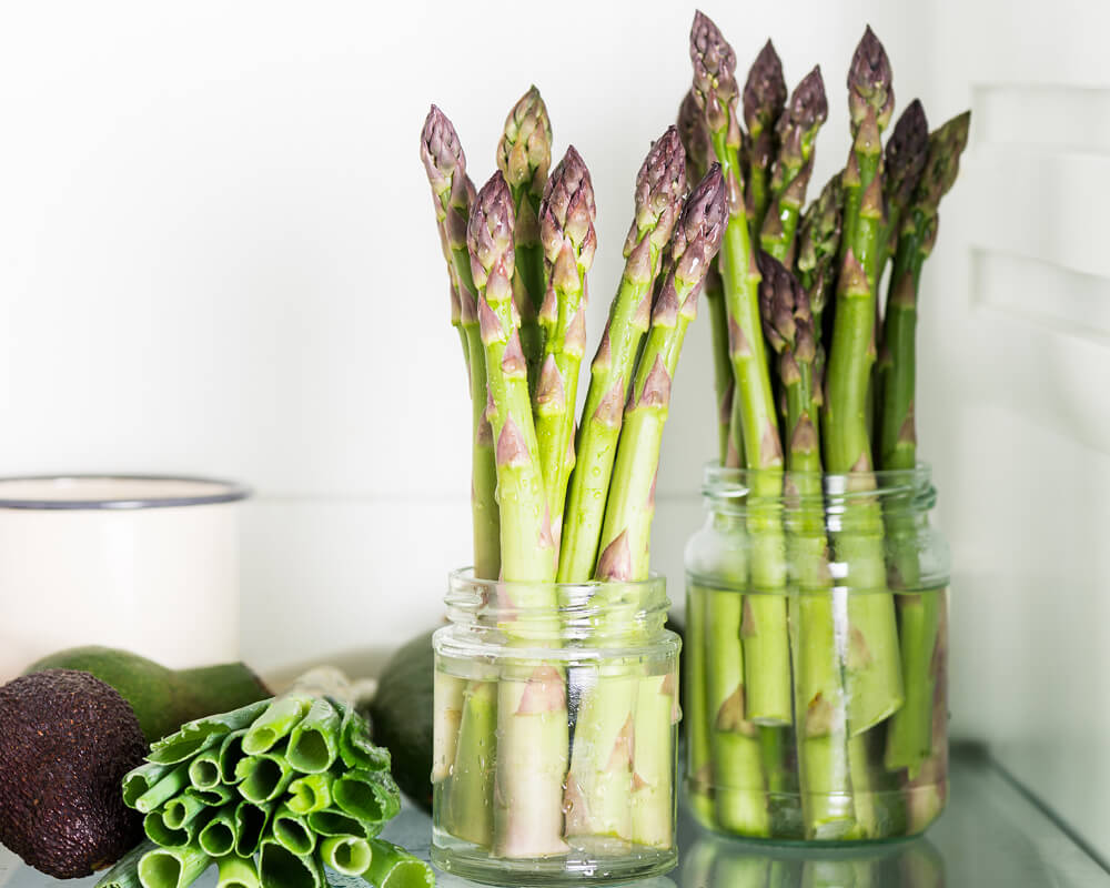 two short glass jars filled with a little water and asparagus stalks sticking upright in them. avocados and a bunch of green onions lay on the glass counter to the left of the jars.