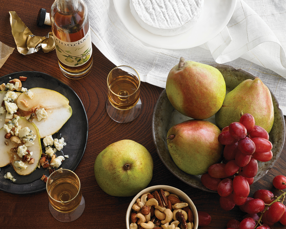 Pears on a table spread with fruit, nuts, cheese, and libations